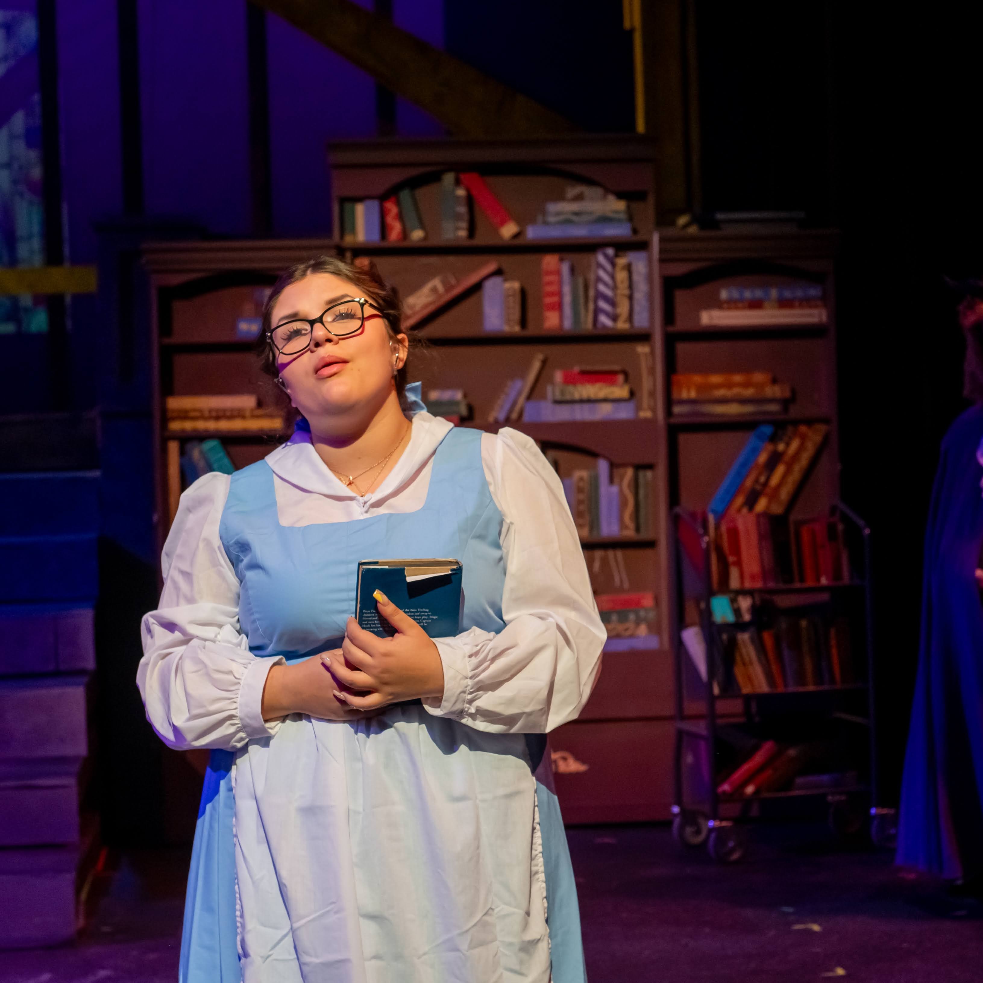 A woman in a blue and white dress stares out towards the skies in our direction. She stands in front of a bookcase.